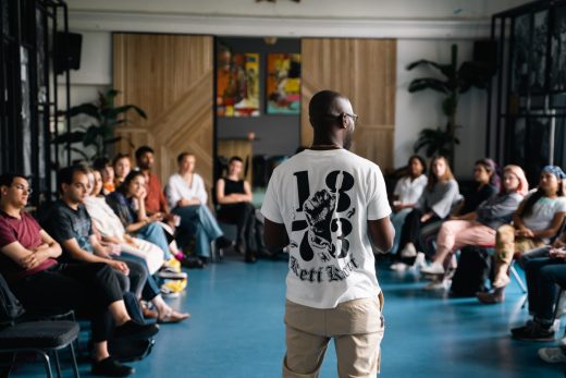 A black man is speaking in front of a group. The photo is taken from behind him and you can see the group sitting in a circle. He is wearing a white t-shirt with numbers on it, he is wearing glasses and is bald.