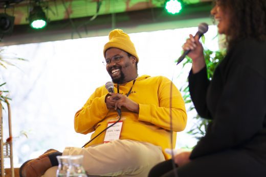 A black man, sitting down, wearing a yellow hoodie and yellow hat, holding a microphone